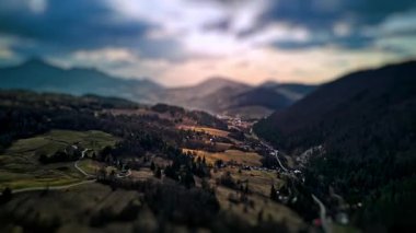 Hareket eden güneş ışınlarıyla yıkanmış bir dağ vadisini gösteren eğik kayma efektiyle sinematik hiperhız. Minyatür görünüm, sarsılan tepeleri, orman kenarlarını ve dramatik bir manzaranın altında huzurlu bir kırsal kasabayı ön plana çıkarıyor.