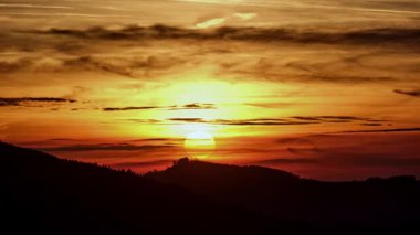 Zaman ayarlı bir video, bir dağ sırtı siluetinin üzerinde çarpıcı bir günbatımını yakalar. Güneşin son ışınları bulutlu bir gökyüzünde turuncu ve kırmızı parlak bir palet oluşturarak nefes kesici ve dramatik bir hava yaratır.
