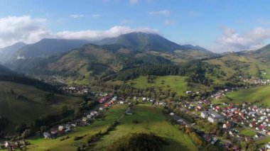 Yeşil tepeler ve ormanlarla çevrili dağınık köy evleri olan bir dağ vadisinin gündüz hava görüntüsü. Parlak güneş ışığı ve yumuşak bulutlar Karpatların barışçıl kırsal atmosferini gözler önüne seriyor.