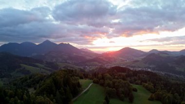 Gün batımında yeşil bir dağ vadisinde geriye doğru uçan hava görüntüleri. Güneş uzak tepelerin ardında batarken dramatik bulutlar gökyüzünde hareket ederek dinamik ve atmosferik bir görünüm yaratıyor.
