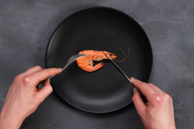 Gri üzerine siyah tabakta büyük bir karides. Sağlıklı deniz ürünleri. Minimalizm, keto zayıflama diyet konsepti. Üst Manzara. Çatal ve bıçak tutan adam elleri.