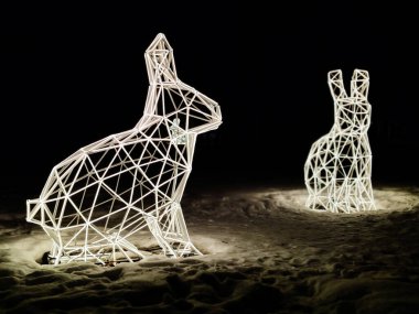 Geceleri karların üzerinde parlayan kablo lambasından yapılmış aydınlatılmış geometrik tavşan heykelleri. Modern tasarımı ve şenlikli atmosferi olan yaratıcı açık hava ışık kurulumu. Kış dekorasyonu, tatil ışıkları, sanat kurulumu kavramı