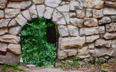 Engebeli, düzensiz taş bloklardan oluşan kalın bir duvarda açılan küçük kemerli bir resmin odaklanmış görüntüsü. Kemerden yemyeşil yapraklar görülebiliyor, tarih ve gizli doğa hissi yaratıyor. Arkeoloji ve antik temalar için mükemmel..