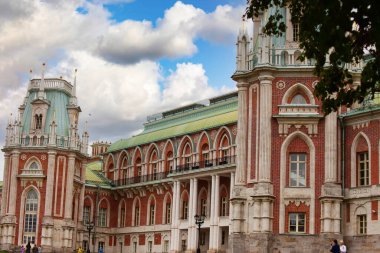 Muhteşem bir saray kompleksinin çarpıcı manzarası. Karmaşık Gotik yeniden canlandırma mimarisi. Ön cephe kırmızı tuğla, beyaz taş aksanlar ve sivri kuleleri olan dekoratif kulelerle karakterize edilir, dinamik bir gökyüzüne karşı kurulur..