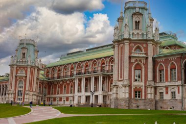 Muhteşem bir saray kompleksinin çarpıcı manzarası. Karmaşık Gotik yeniden canlandırma mimarisi. Ön cephe kırmızı tuğla, beyaz taş aksanlar ve sivri kuleleri olan dekoratif kulelerle karakterize edilir, dinamik bir gökyüzüne karşı kurulur..