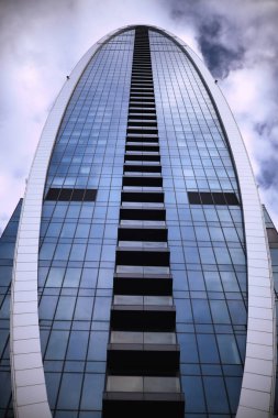 Modern bir cam gökdelenin simetrik, eğri büğrü siluetini yakalayan profesyonel bir mimari fotoğraf. Düşük açılı perspektif, balkonların merkezi dikey çizgisini ve mavi cephenin pürüzsüz, yansıtıcı yüzeyini vurgular..