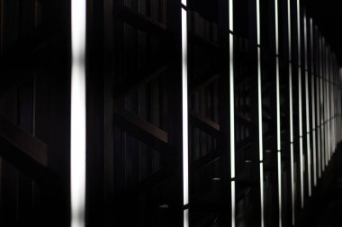 Modern bir mimari tesisin profesyonel gece çekimleri. Görüntü, parlak dikey LED ışıklar tarafından çerçevelenmiş koyu metal zig-zag destekli ritmik geometrik bir desen içermekte olup, yüksek kontrastlı ve fütüristik şehir arkaplanı oluşturmaktadır..