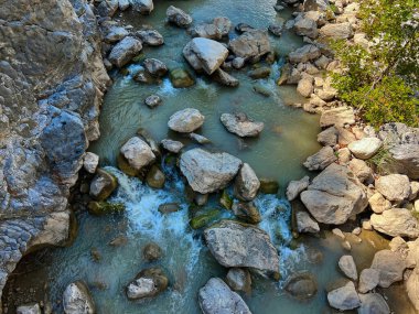 Dinamik bir dağ nehrine bakan yüksek açılı bir çekim. Su, dağınık büyük gri kayalar arasında akar ve küçük beyaz su akıntıları oluşturur. Islak taşların ve etrafındaki kayalıkların doğal dokuları vurgulanmıştır.