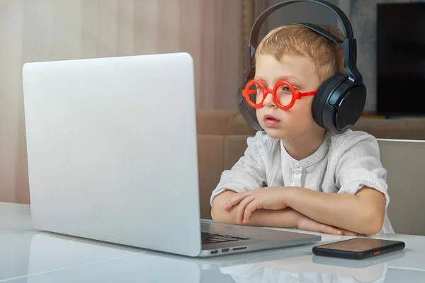 Ένα αγόρι που φοράει ασύρματα ακουστικά κάθεται σε ένα γραφείο χρησιμοποιεί ένα lapto — Φωτογραφία Αρχείου
