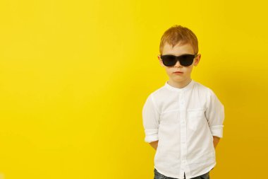 Güneş gözlüklü bir çocuk sarı arka planda poz veriyor.