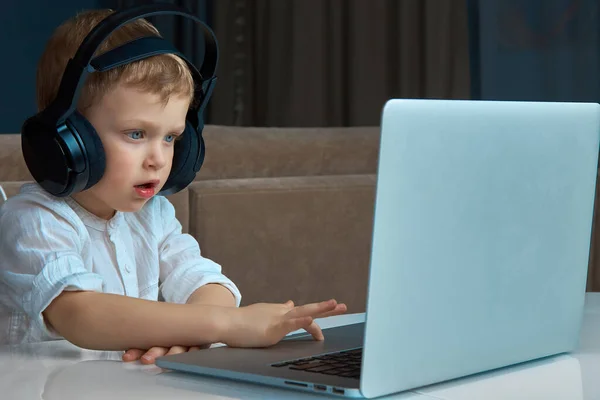 Ένα παιδί με μπλε μάτια που φοράει ακουστικά χρησιμοποιώντας ένα touchpad ελέγχει ένα laptop — Φωτογραφία Αρχείου