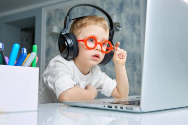 Kırmızı gözlüklü ve siyah kulaklıklı bir çocuk dizüstü bilgisayarı inceliyor.