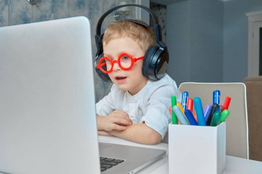Kırmızı gözlüklü ve kulaklıklı bir çocuk bilgisayar kullanarak video bağlantısıyla iletişim kurarken eğleniyor..