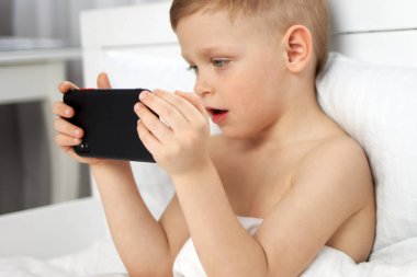 Küçük bir çocuk beyaz bir yatakta telefona dikkatle bakıyor.