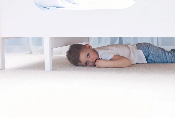 Korkmuş bir çocuk beyaz bir yatağın altında saklanıyor. — Stok fotoğraf