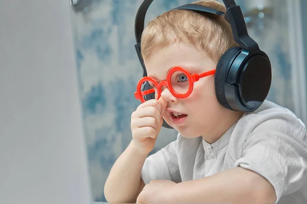 Ένα αγόρι με αστεία κόκκινα γυαλιά εκπαιδεύεται διαδικτυακά σε ένα λάπτοπ και σκαλίζει τη μύτη του από βαρεμάρα. — Φωτογραφία Αρχείου