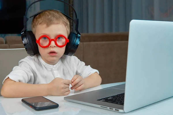 Ένα αγόρι με μπλε μάτια και γυαλιά χρησιμοποιεί φορητό υπολογιστή και ασύρματα ακουστικά — Φωτογραφία Αρχείου