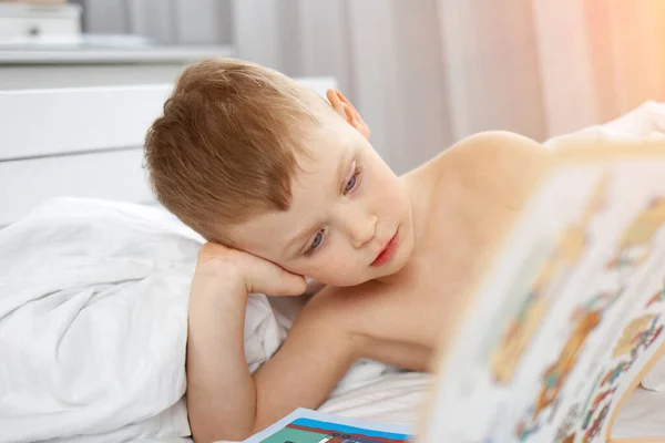 Aranyos fiú fekszik egy fehér ágyban, és egy varázskönyvet olvas. — Stock Fotó