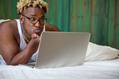 Siyah adam, modern iletişim araçlarını kullanarak online olarak yatakta yatıyor. Genç bir Afro-Amerikalı dizüstü bilgisayara öpücük yolluyor.