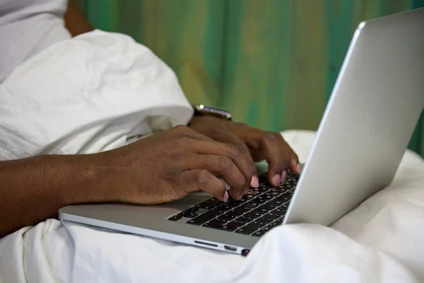 Close-up of a black African man typing with his hands on a laptop computer sitting on a white bed