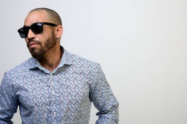 Sakallı, güneş gözlüklü ve beyaz arka planda gömleği olan havalı bir Latin adamın portresi.