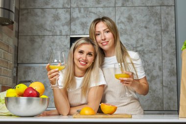 İki tatlı sarışın kadın mutfaktaki bardaklardan portakal alkollü kokteyl içiyor ve taze meyve yiyorlar.
