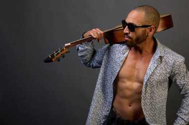 Güneş gözlüklü, kaslı Latin Amerikalı ve gri arka planda omzunda akustik gitar tutan bir gömlek.