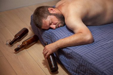 Sarhoş bir adam elinde bira şişesiyle evde bir yatakta uyuyor. Yanında boş şişeler var..