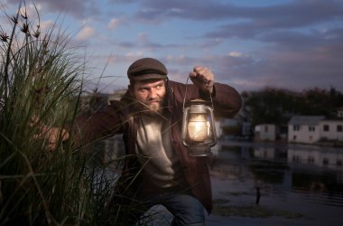 Kötü yüzlü, sakallı ve klasik elbiseli bir adam elinde bir gaz lambası tutuyor. Etrafı karanlık, deniz arkası.