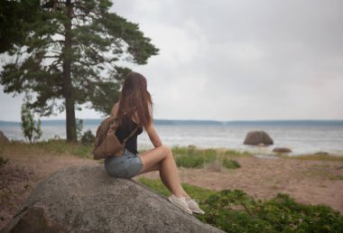 Sırt çantalı bir kız deniz kıyısındaki bir ağacın altında bir kayanın üzerinde oturuyor. Arkadan bak.