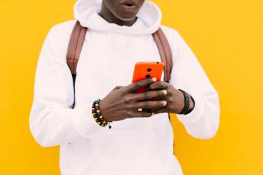 Genç Afrikalı bir adam sarı bir duvara yaslanmış beyaz bir kazak ve kablosuz kulaklıkla müzik dinleyen bir sırt çantası giyiyor.