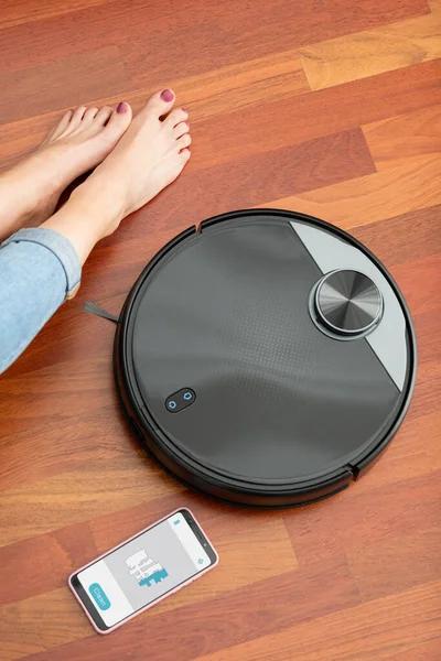 Akıllı telefonu ve yerde otomatik elektrikli süpürgesi olan genç bir kadının yüzü olmayan orta kesimi. Uygulamalı ev işi robotu