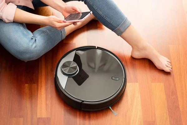 Yerleri temizlemek için elektrikli süpürge kullanan, akıllı ev işi robotu akıllı telefonla kontrol eden genç bir kadının yüzsüz orta bölümü.