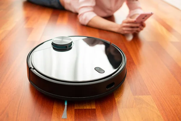 Yerleri temizlemek için elektrikli süpürge kullanan, akıllı ev işi robotu akıllı telefonuyla kontrol eden genç bir kadının yüzsüz orta bölümü.