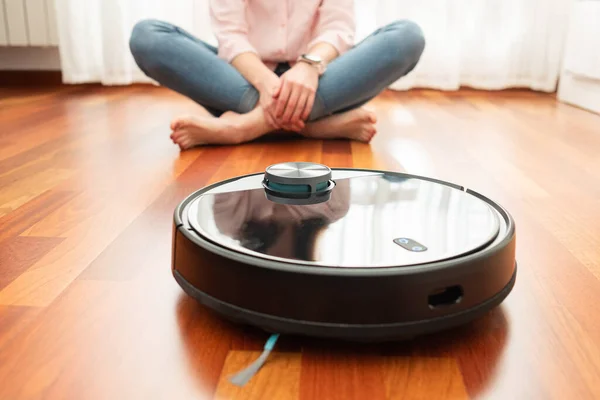 Faceless middle section of young woman seated at floor while using automatic vacuum cleaner to clean the floor. Future Technology, internet of things iot concept