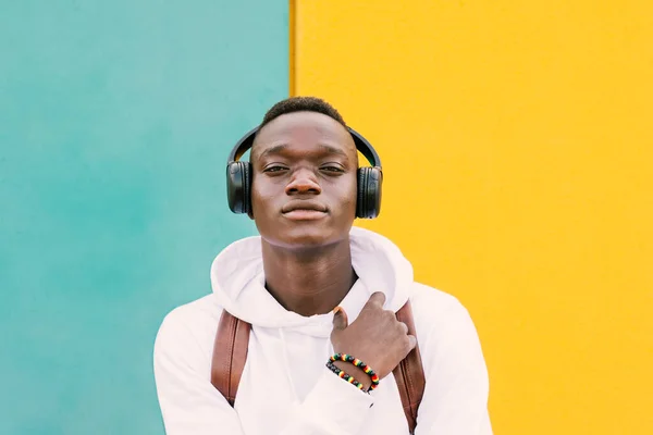 Genç Afrikalı bir adam sarı ve yeşil bir duvara karşı beyaz bir kazak ve kablosuz kulaklıkla müzik dinleyen bir sırt çantası giyerek kameraya bakıyor.