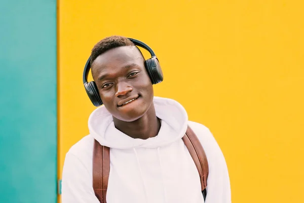 Beyaz eşofman üstü ve sırt çantası takmış, yeşil ve sarı arka planda kameraya bakarken kablosuz kulaklıkla müzik dinleyen Afro Amerikalı genç bir adam.