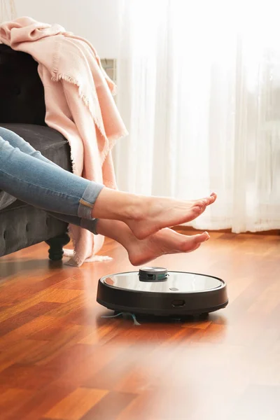 Genç bir kadının, koçun üzerinde dinlenen yüzü olmayan orta bölümü yerleri otomatik süpürge kullanarak temizliyor, akıllı makine ev işi robotunu kontrol ediyor.