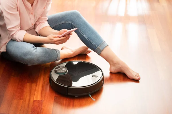 Yerleri temizlemek için elektrikli süpürge kullanan, akıllı ev işi robotu akıllı telefonla kontrol eden genç bir kadının yüzsüz orta bölümü.