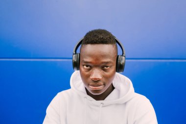 Beyaz bir kazak giyerken kablosuz kulaklıkla müzik dinleyen Afro Amerikalı genç adam üzgün bir ifadeyle kameraya bakıyor.