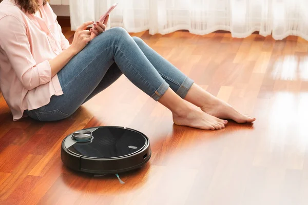 Yerleri temizlemek için elektrikli süpürge kullanan, akıllı ev işi robotu akıllı telefonla kontrol eden genç bir kadının yüzsüz orta bölümü.