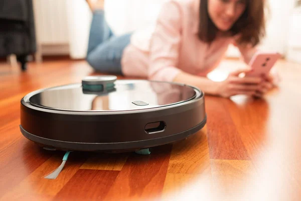 Yerleri temizlemek için elektrikli süpürge kullanan, akıllı ev işi robotu akıllı telefonuyla kontrol eden genç bir kadının yüzsüz orta bölümü.