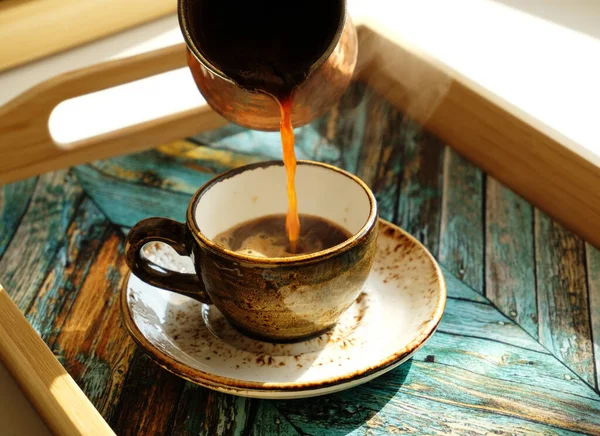 Café turco caliente vertiendo desde el cezve en una taza de café expreso de cerámica en un platillo. Luz del día, sombras. Bandeja de madera turquesa — Foto de Stock