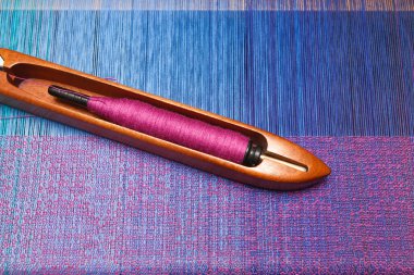 Weaving shuttle with thread on the blue warp clipart