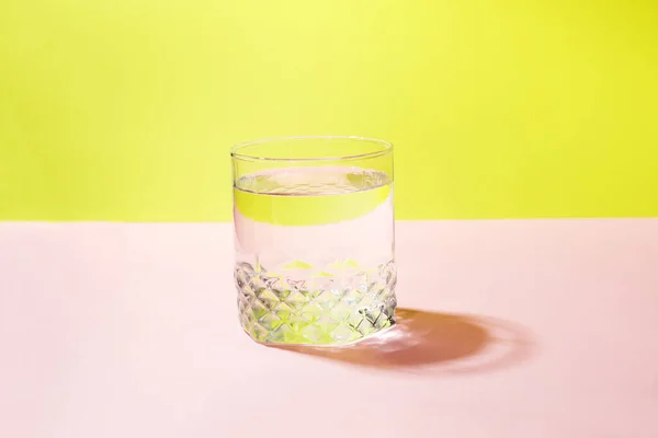 Renkli pembe ve sarı arka planda saf ferahlatıcı berrak bir su bardağı. Vücut sağlığı için günlük su tüketimi kavramı