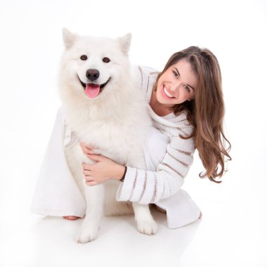 Beyaz köpeğiyle birlikte beyaz giyinmiş, poz veren, mutlu görünen ve gülümseyen genç bir kadının stüdyo görüntüsü.