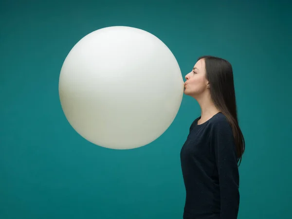 Uzun siyah saçlı komik genç kadın mavi arka planda kocaman bir balon şişiriyor.