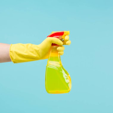 Elinde lastik eldiven, açık mavi arka planda sarı temizlik spreyi şişesi var.