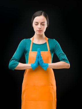 Önlük ve lastik eldiven giyen genç temizlikçi kadın siyah arka plana karşı dua ediyor ya da meditasyon yapıyor.