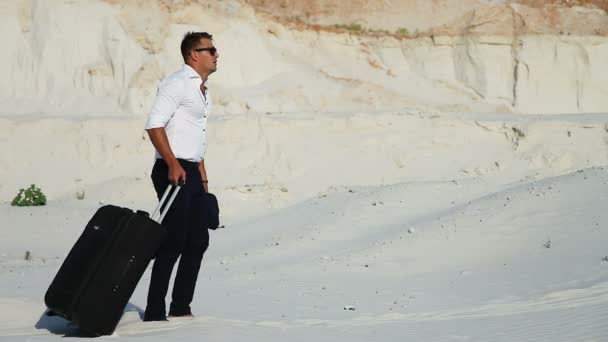 Geschäftsmann in der Wüste. Ein junger Mann in Businessanzug und großer Gepäcktasche in der Wüste. Geschäftsmann auf dem Weg zum Ferienort. weißer Sand. Schwüle Wüste. — Stockvideo